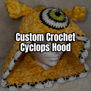May include: A custom crochet cyclops hood in yellow with a large eye design. The eye is white, black, and green. The hood has two small horns and a white and black trim. The text "Custom Crochet Cyclops Hood" is displayed on the hood.