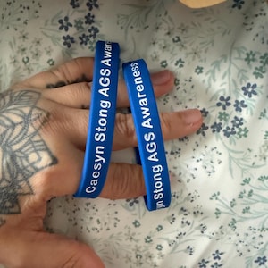 May include: Two blue silicone wristbands featuring white text that reads Caesyn Stong AGS Awareness. The bracelets are flexible and designed to be worn around the wrist, displaying a clear message of support for a specific cause or individual.