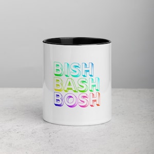 Puede incluir: Taza de cerámica blanca con interior negro. La taza presenta las palabras "BISH BASH BOSH" apiladas verticalmente en una fuente de gradiente de arcoíris. La taza está sobre un fondo neutro.