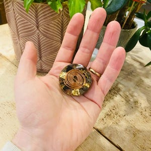 以下が含まれることがあります： 丸い手作りのオルゴンエネルギーデバイス。デバイスは、中央に銅コイル、金箔と濃い樹脂で囲まれています。デバイスは開いた手で保持され、指には金の指輪が付いています。