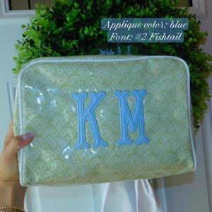May include: A rectangular zippered pouch featuring a light green and white fan-shaped pattern with a white piped edge. The center displays the blue embroidered monogram letters KM in a fishtail font. Text on the image reads: Applique color: blue, Font: #2 Fishtail.