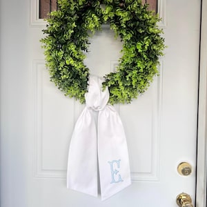 May include: A circular wreath composed of dense green boxwood foliage, featuring a long, white fabric ribbon tied at the bottom. The ribbon hangs in two tails, with the letter E embroidered in light blue thread on the lower right side of the fabric.