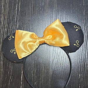 May include: Black headband with two round ears featuring a gold glasses pattern. A large, shiny gold bow is centered between the ears. The headband is a fun accessory for themed events or everyday wear.
