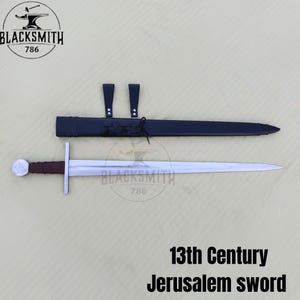 May include: A 13th Century Jerusalem sword with a silver-toned pommel, a dark brown wrapped grip, and a straight crossguard. The blade is metallic silver. It is accompanied by a black leather scabbard featuring two vertical straps and a logo.