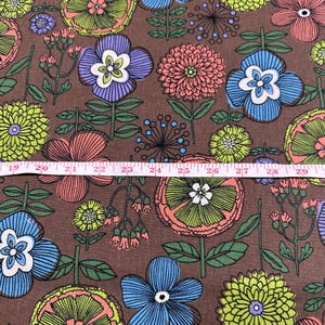 May include: Brown floral fabric with a repeating pattern of stylized flowers in shades of blue, purple, orange, and green. The flowers have detailed petals and are set against a brown background. A measuring tape is laid across the fabric.