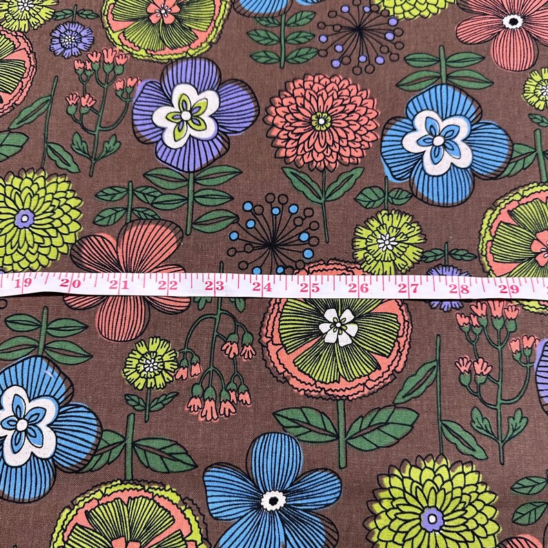 May include: Brown floral fabric with a repeating pattern of stylized flowers in shades of blue, purple, orange, and green. The flowers have detailed petals and are set against a brown background. A measuring tape is laid across the fabric.