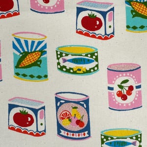 May include: A cream-colored fabric featuring a repeating pattern of various canned goods. The illustrations include cans of tomatoes, corn, fish, and cherries, rendered in vibrant colors like red, blue, green, and pink against a neutral background.
