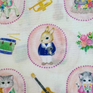 May include: A light-colored fabric featuring a repeating pattern of circular frames. Inside the circles are illustrations of a rabbit in a blue jacket holding a small toy, kittens, and floral arrangements. Musical instruments like a trumpet and drum are also visible.