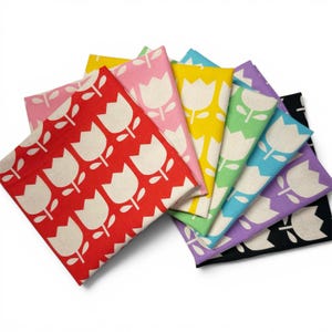 May include: A collection of square fabric napkins featuring a repeating white tulip pattern. The set includes napkins in red, light pink, yellow, light green, light blue, purple, and black, all arranged in a fanned-out, overlapping stack.