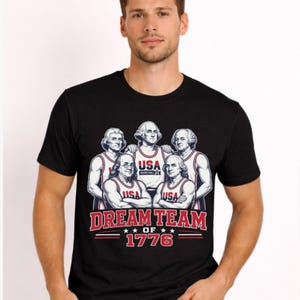 Può includere: Una t-shirt nera a maniche corte con una grafica raffigurante cinque personaggi storici che indossano maglie da basket USA. Sotto le figure, il testo recita DREAM TEAM OF 1776 in lettere rosse e bianche, con due stelle su ogni lato della parola OF.