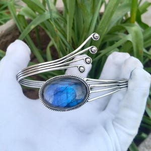 Può includere: Un bracciale a fascia in argento con una pietra preziosa di labradorite ovale centrale dai riflessi blu. Il bracciale è composto da diversi fili d'argento sottili che si aprono a ventaglio a un'estremità, terminando con una piccola perla.