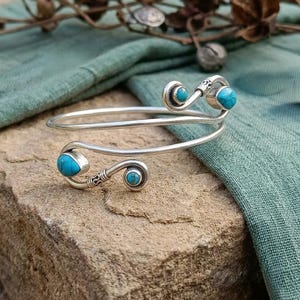May include: A silver-toned bypass cuff bracelet featuring a spiral design at each end. The bracelet is accented with four small, round and teardrop-shaped turquoise stones set into the metal, with decorative etched detailing near the stone settings.