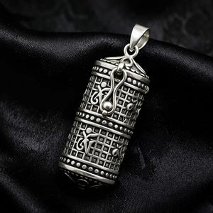 Könnte beinhalten: Ein silberner Anhänger mit einem verzierten, zylindrischen Design. Der Anhänger weist komplizierte Muster auf, darunter geometrische Formen und dekorative Elemente. Er hat eine Öse zum Anbringen an einer Kette und einen Verschluss. Der Anhänger wird vor einem dunklen Hintergrund präsentiert.