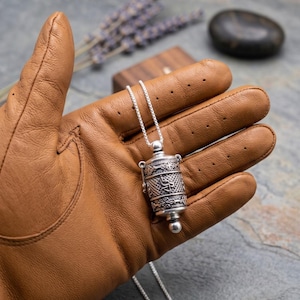 May include: A silver pendant necklace featuring a detailed cylindrical charm with intricate patterns. The charm hangs from a delicate silver chain. The pendant is held in a brown leather glove, with a blurred background of lavender and a dark stone.