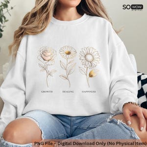 Op de afbeelding: Witte sweatshirt met een gouden lijntekening van een roos, madeliefje en zonnebloem. De woorden "GROWTH", "HEALING" en "HAPPINESS" staan onder de bloemen. De tekst "SO WOW DIGITAL" staat rechtsboven.