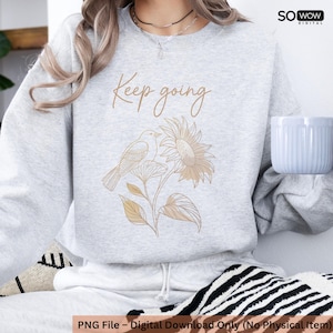Puede incluir: Una sudadera gris claro con la frase "Keep going" en fuente cursiva sobre una ilustración de un pájaro y un girasol. El pájaro y la flor son de color marrón claro. Una taza blanca se sostiene en la mano derecha.