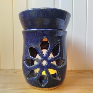 Könnte beinhalten: Ein dunkelblauer Keramik-Diffusor für ätherische Öle mit einem blütenförmigen Ausschnitt. Der obere Teil hat eine Schalenform. Im Inneren ist eine brennende Kerze zu sehen, die ein warmes Licht wirft. Der Diffusor steht auf einer Holzoberfläche.