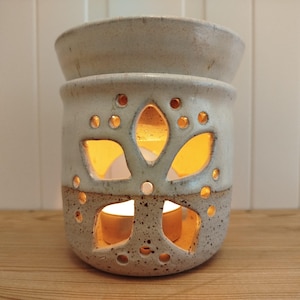 Könnte beinhalten: Ein ätherisches Öl Diffusor aus Keramik mit einer hellbeigen, gesprenkelten Oberfläche. Der Diffusor hat ein florales Ausschnittdesign mit kleinen kreisförmigen Löchern und größeren blütenblattförmigen Öffnungen. Eine brennende Kerze im Inneren wirft einen warmen Schein.