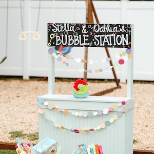 Può includere: Un chiosco di bolle blu chiaro con un cartello che recita "Stella & Dahlia's Bubble Station". Il chiosco ha ghirlande di cerchi di carta colorati e due secchi pieni di giocattoli a bolle. Una macchina per bolle rossa e verde è appoggiata sul bancone.