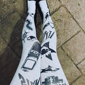 May include: White tights patterned with black graphics, including a centipede, eye, starburst, and a flip phone. The tights also feature text and images of a shark and other shapes. The tights are worn over black shoes.