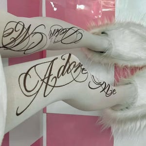 May include: White tights with black cursive text that reads "Adore Me" and "Dear". The tights are paired with white fluffy slippers. The background is a pink and white checkered pattern.