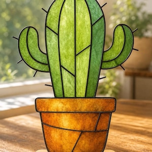 May include: A decorative cactus ornament featuring green translucent panels framed in black metal, set in an amber-coloured pot with a segmented design. The cactus has two side arms with small metal spikes and rests on a rectangular wooden base.