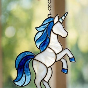 May include: A stained glass unicorn suncatcher featuring a white body with a flowing blue mane and tail. The unicorn is depicted in a rearing pose with a single horn, constructed with dark metal lead lines and suspended by a silver metal chain.