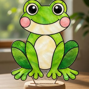 May include: A stained glass frog figure featuring vibrant green glass panels, a pale yellow belly, and soft pink circular cheeks. The frog has large black eyes with white highlights and is mounted on a smooth, circular light-coloured wooden base.