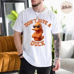 May include: White t-shirt featuring a cartoon duck wearing sunglasses and an orange hoodie. The text "DON'T GIVE A DUCK" is printed above and below the duck graphic in orange block letters. The shirt is a casual style.