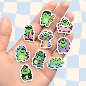May include: A collection of nine small die-cut stickers featuring a green frog in various outfits and activities, including a frog with a baguette, in a bathtub, on a scooter, in a UFO, wearing a hoodie, a dinosaur costume, a cap, and a leather jacket.