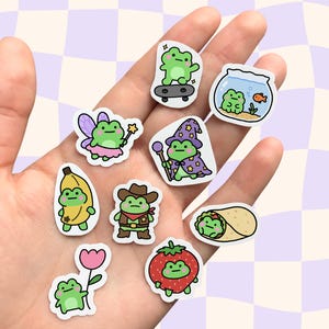 May include: A collection of nine small die-cut stickers featuring green frogs in various costumes and scenarios, including a frog on a skateboard, in a fishbowl, as a fairy, a wizard, a banana, a cowboy, a burrito, with a flower, and as a strawberry.