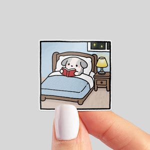 May include: A sticker featuring a cartoon dog in bed reading a book. The dog is under a blue and white checkered blanket, with a bedside table and lamp. The background shows a window with stars.