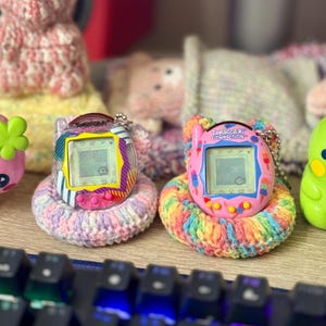 Könnte beinhalten: Zwei Tamagotchi Connection Digital-Haustiere auf gehäkelten Garnringen. Das linke Gerät hat ein Gehäuse mit Streifen und geometrischen Mustern, während das rechte Gerät rosa ist, mit bunten Eiscreme-Grafiken und einem digitalen Bildschirm.