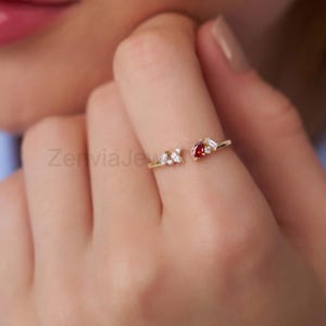 May include: A gold-colored ring with an open design, featuring a cluster of small white stones and a pear-shaped red stone. The ring is worn on a finger, with the brand name "ZerviaJewels" visible in the background.