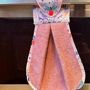 Pode incluir: Um pano de cozinha rosa com estampa floral e borboleta, pendurado em uma barra de metal. A toalha tem um topo decorativo com um botão vermelho e acabamento em renda branca. A toalha é feita de um material macio e absorvente.