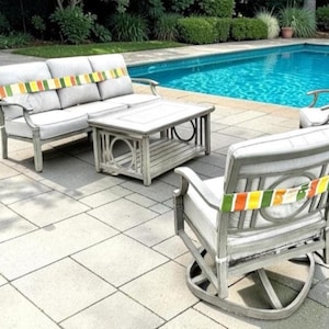 May include: Outdoor patio furniture set with a light gray finish. The set includes a three-seat sofa, two swivel chairs, and a coffee table. The cushions are white, and the backs of the chairs and sofa have a colorful striped accent.