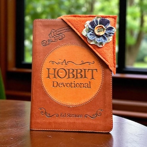 May include: A brown leather-bound book titled "A Hobbit Devotional" with an orange felt bookmark featuring a blue and green fabric flower with a button center. The book is on a wooden table.