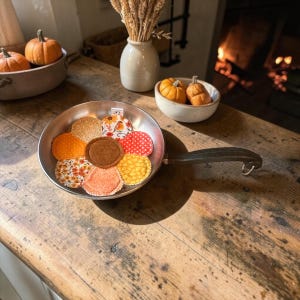 May include: A metal frying pan containing several circular fabric coasters arranged in a flower pattern. The coasters feature various autumn-themed prints including orange, yellow, and red patterns, with a solid brown center piece.