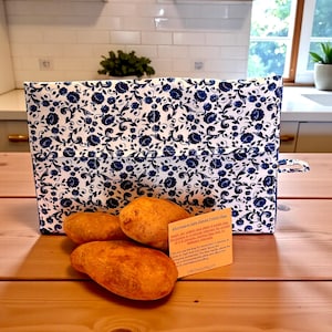 May include: A white and blue floral patterned microwave baked potato bag with three baked potatoes on a wooden surface. A small card with text is visible. The bag has a handle on the side.