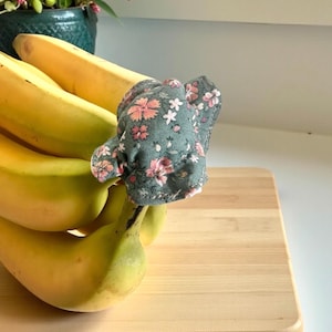 Könnte beinhalten: Ein Bund reifer gelber Bananen mit einem Bananenschutz aus floralem Stoff. Der Schutz ist in gedämpftem Grün mit rosa und weißen Blumenmustern gehalten. Die Bananen liegen auf einem hellen Holzschneidebrett.
