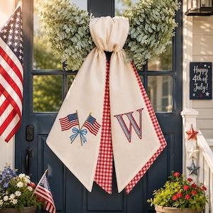May include: Cream-colored door scarf with red gingham trim and embroidered American flags and the letter "W". The scarf hangs on a blue door with a wreath and a sign that says "Happy 4th of July!"