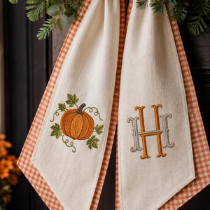 May include: A cream-coloured fabric hanging decoration featuring an orange pumpkin with green leaves embroidered on the left side and a large, stylised orange and grey letter H monogram embroidered on the right side, finished with orange gingham trim.