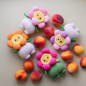 Op de afbeelding: Een bovenaanzicht toont verschillende handgemaakte gehaakte bloemenspeeltjes in roze, oranje en paars, elk met een lachend gezicht. De speeltjes worden gecombineerd met verse perziken, wat zorgt voor een kleurrijk en speels arrangement.