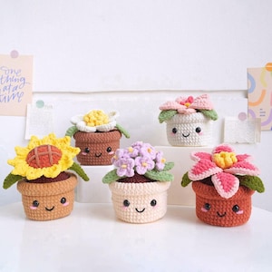 Puede incluir: Una colección de cinco decoraciones de macetas de ganchillo. Cada maceta tiene una cara sonriente y está llena de una flor de ganchillo diferente, incluyendo un girasol, una flor blanca, una flor rosa, una flor morada y una flor roja y rosa. Las macetas son de varios colores.