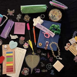 Puede incluir: Una colección de artículos de papelería y accesorios, que incluye un cuaderno azul, bolígrafos, pegatinas, un estuche y un collar de cuentas. Los artículos están dispuestos sobre una superficie negra, con una variedad de colores y texturas.