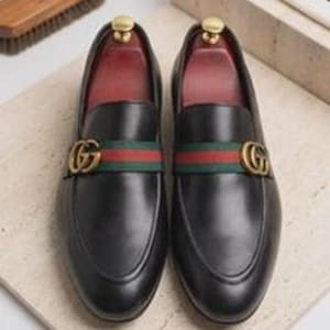 May include: A pair of black leather loafers featuring a green and red fabric stripe across the vamp, accented with a gold-toned metal GG logo emblem. The interior of the shoes is lined with a deep red material, and each shoe contains a wooden shoe tree.