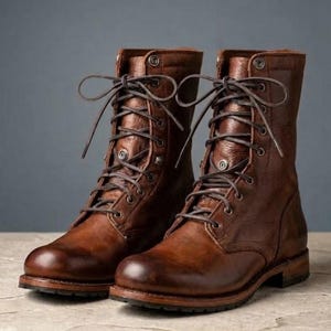Puede incluir: Un par de botas con cordones a media pantorrilla en cuero marrón con acabado desgastado. Estas botas cuentan con cordones marrón oscuro, ojales metálicos plateados y un tacón con suela de goma resistente, ofreciendo una silueta estructurada.