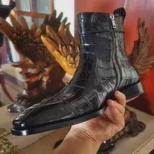 Op de afbeelding: Een zwarte enkelboot met een glanzende afwerking en een textuur van krokodillenleer. De laars heeft een puntige neus, een lage blokhak en een verticale zilverkleurige metalen ritssluiting aan de zijkant van de schacht bij de enkel.