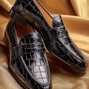 May include: A pair of black loafers featuring a glossy, embossed crocodile skin texture. The shoes have a classic slip-on design with a rounded toe, a low heel, and a stitched apron front detail, showcasing a polished finish and structured silhouette.