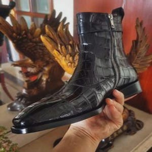Può includere: Uno stivaletto nero con finitura lucida e motivo testurizzato in pelle di coccodrillo. Lo stivale presenta una punta affusolata, un tacco basso a blocco e una cerniera in metallo color argento che corre verticalmente lungo il lato.